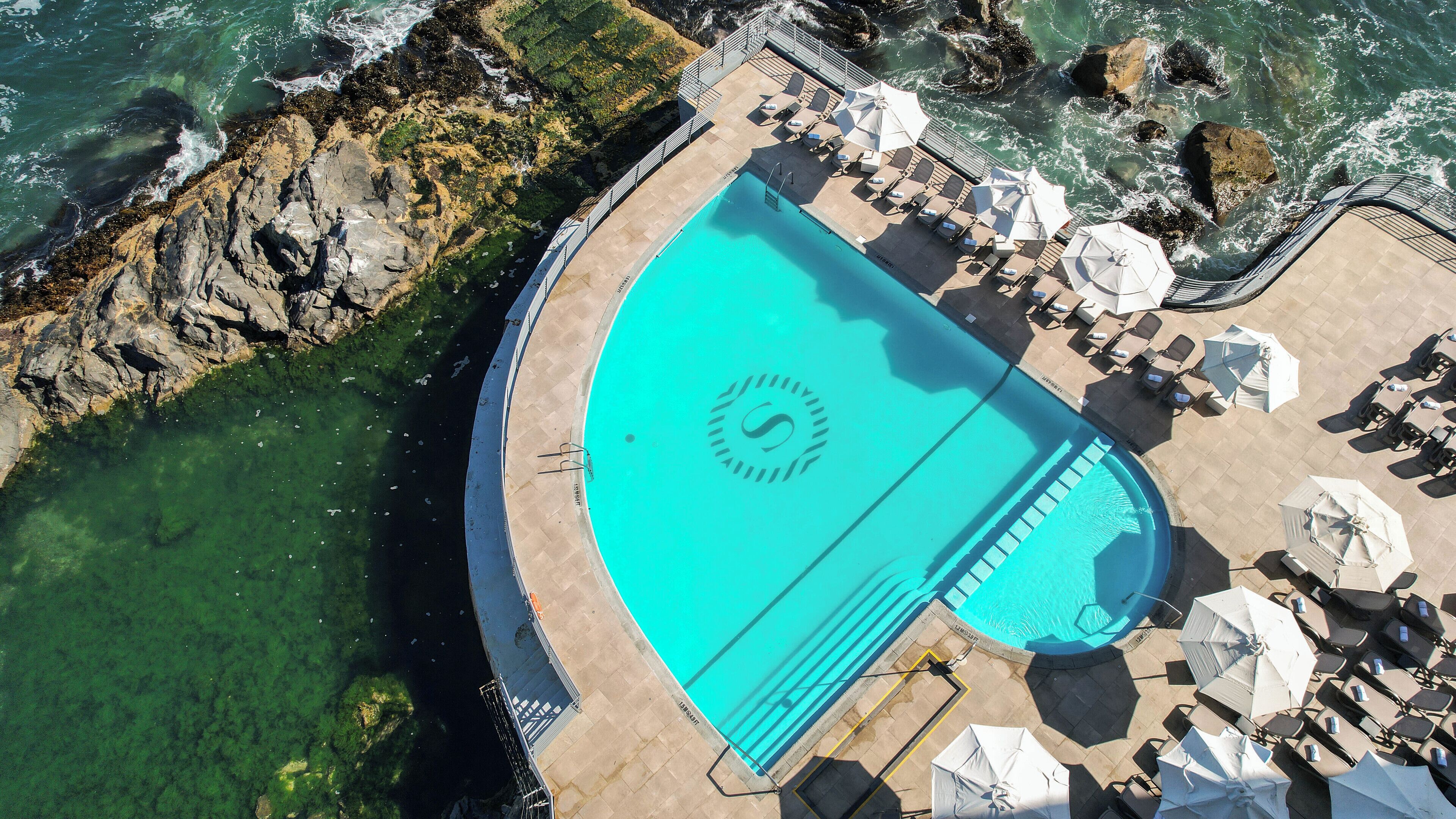 An aerial view of a pool with a logo, surrounded by lounge chairs and umbrellas, with rocky coastline nearby.