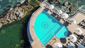 An aerial view of a pool with a logo, surrounded by lounge chairs and umbrellas, with rocky coastline nearby.