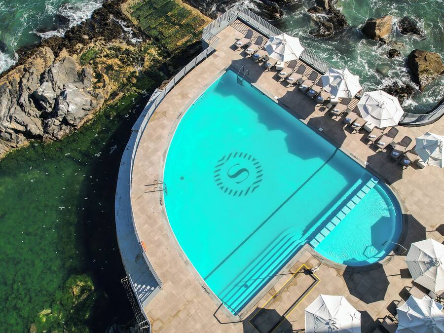 An aerial view of a pool with a logo, surrounded by lounge chairs and umbrellas, with rocky coastline nearby.