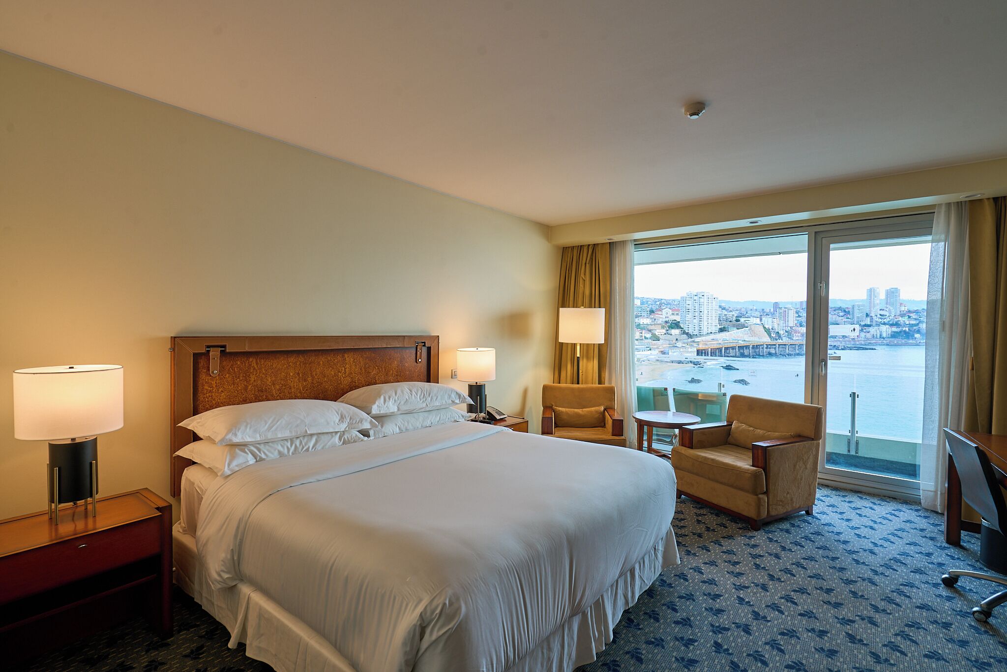 Habitación de hotel con una cama grande, dos lámparas de noche, una silla, una mesita y vistas a un paisaje urbano a través de ventanas amplias.