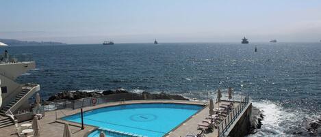 Zona de piscina con sillas de descanso y sombrillas, con vista al mar y barcos a lo lejos.
