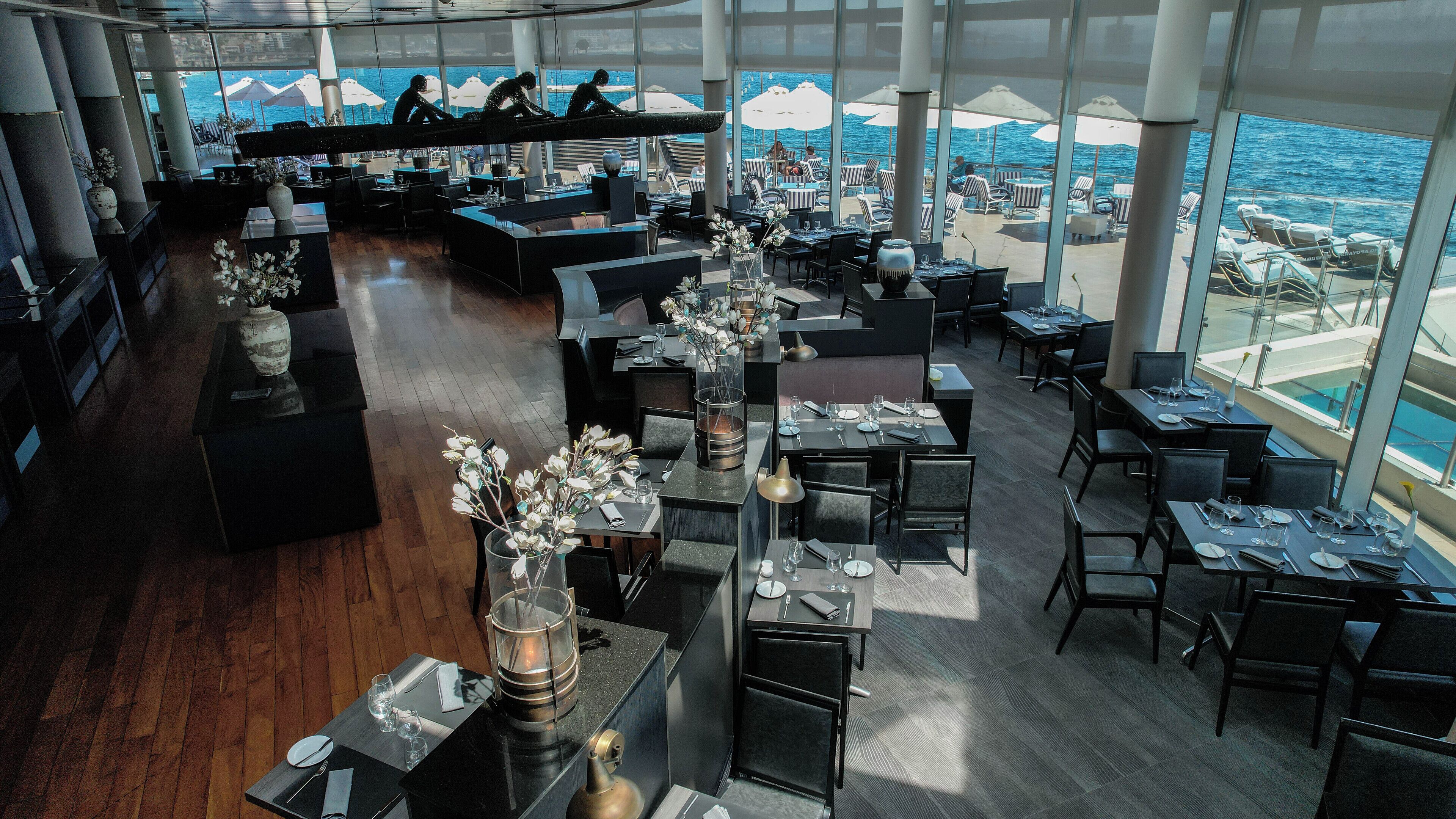 A restaurant with a view of the ocean, featuring tables with chairs, a bar area, and large windows.