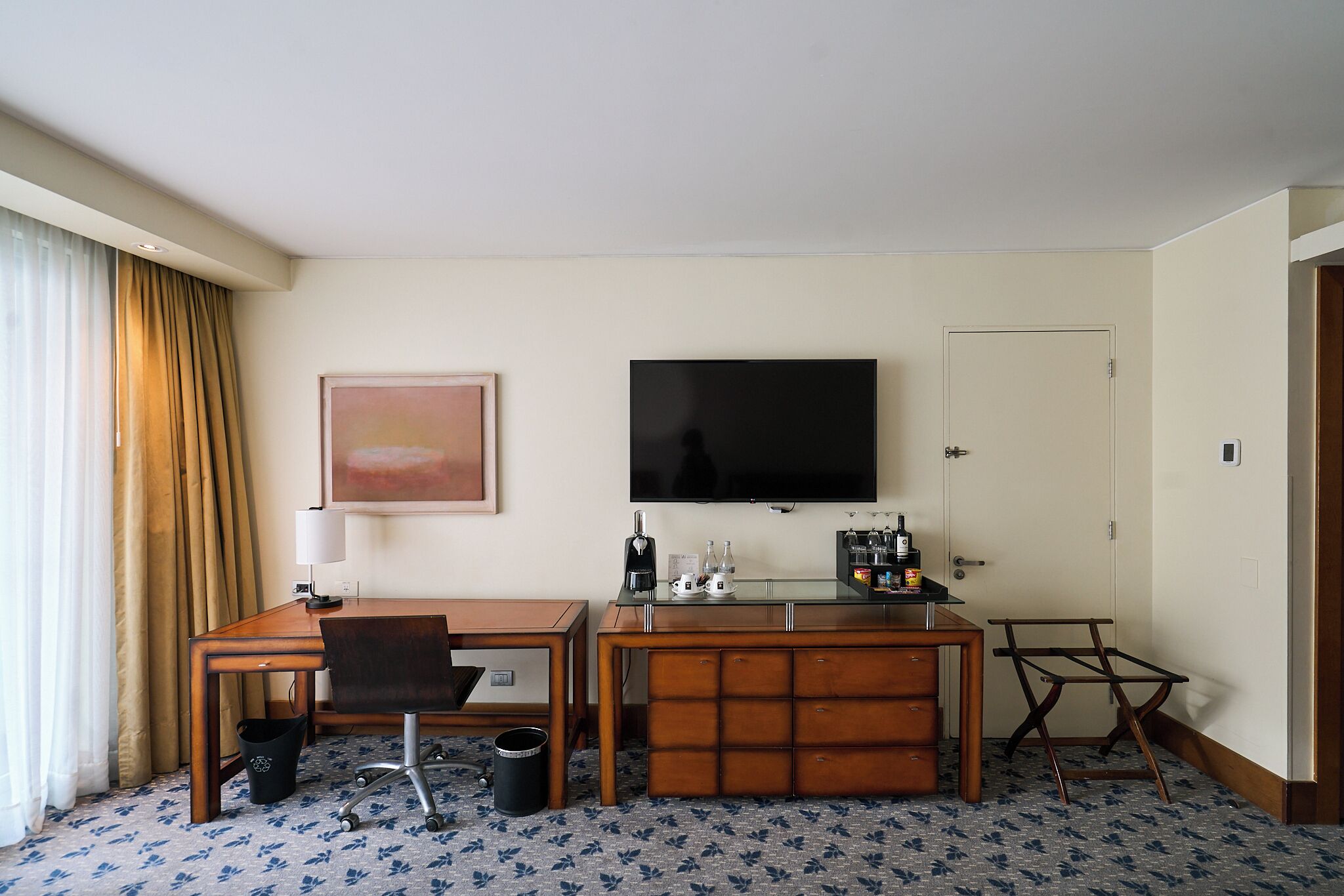 A hotel room with a wooden desk, a television, a minibar, and a painting on the wall.