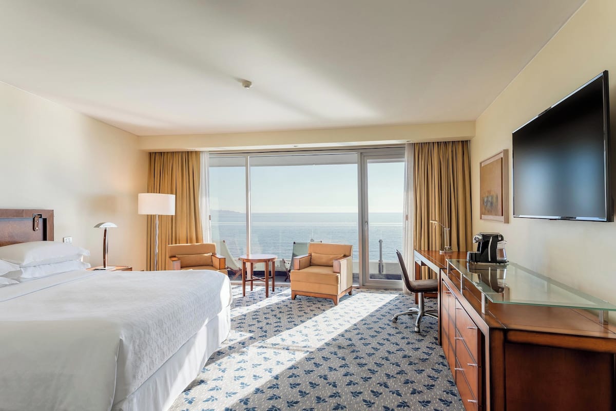 Habitación de hotel con cama, escritorio, silla, lámpara y un amplio ventanal con vista al mar.