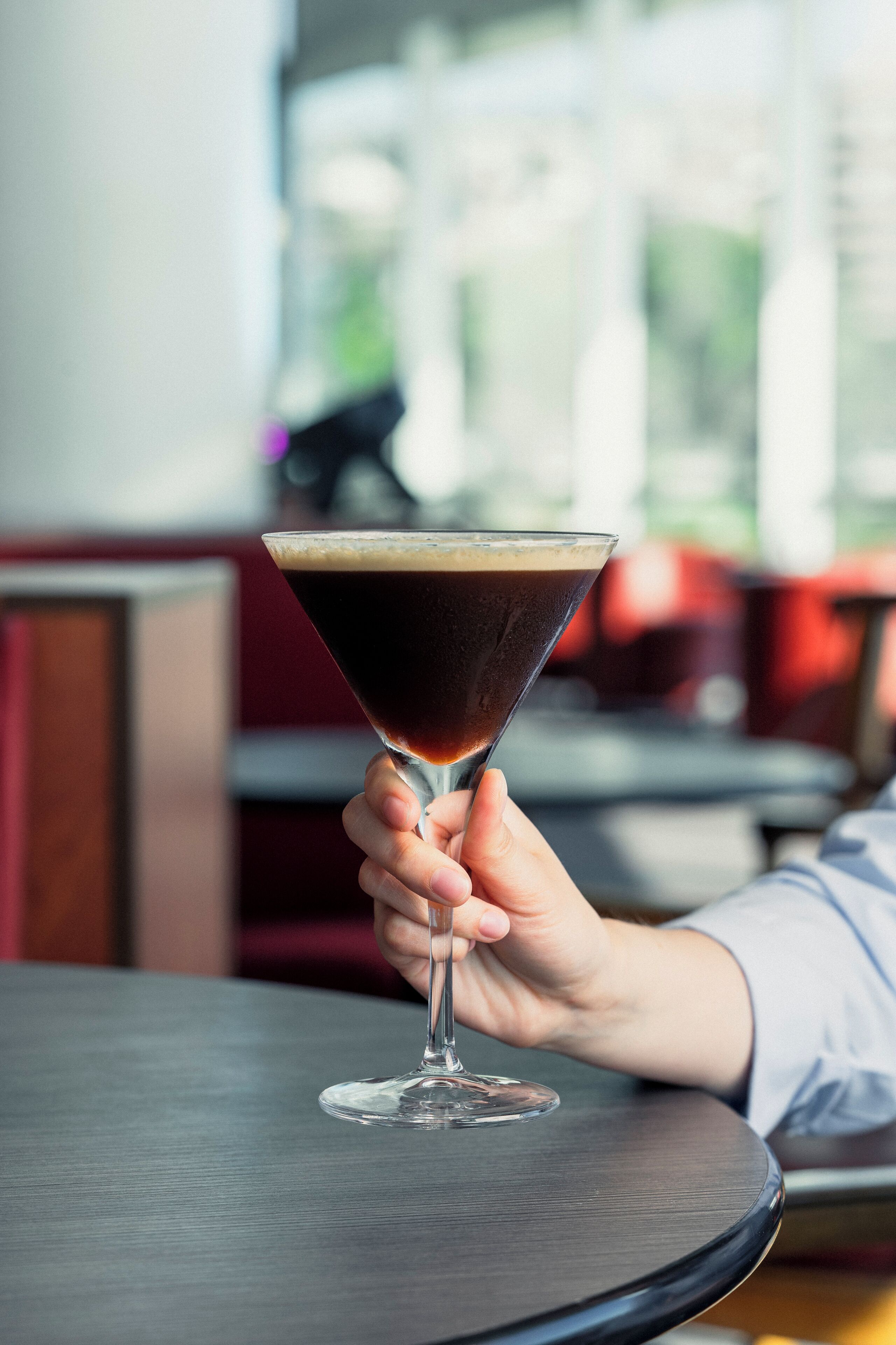 Una mano sosteniendo una copa de martini con un líquido oscuro sobre una mesa redonda.