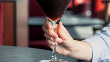 Una mano sosteniendo una copa de martini con un líquido oscuro sobre una mesa redonda.