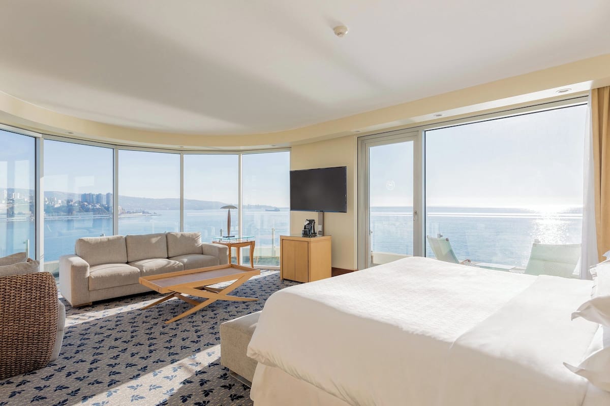 Habitación de hotel con una cama grande, un sofá, una mesa de centro, un televisor y vistas al mar.