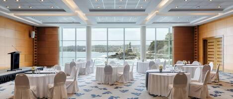A conference room with round tables and chairs, large windows with a view of the sea, and a stage with a podium.
