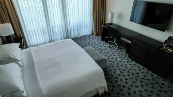 Habitación de hotel con una cama grande, un escritorio, una silla, un televisor y un ventanal con cortinas.