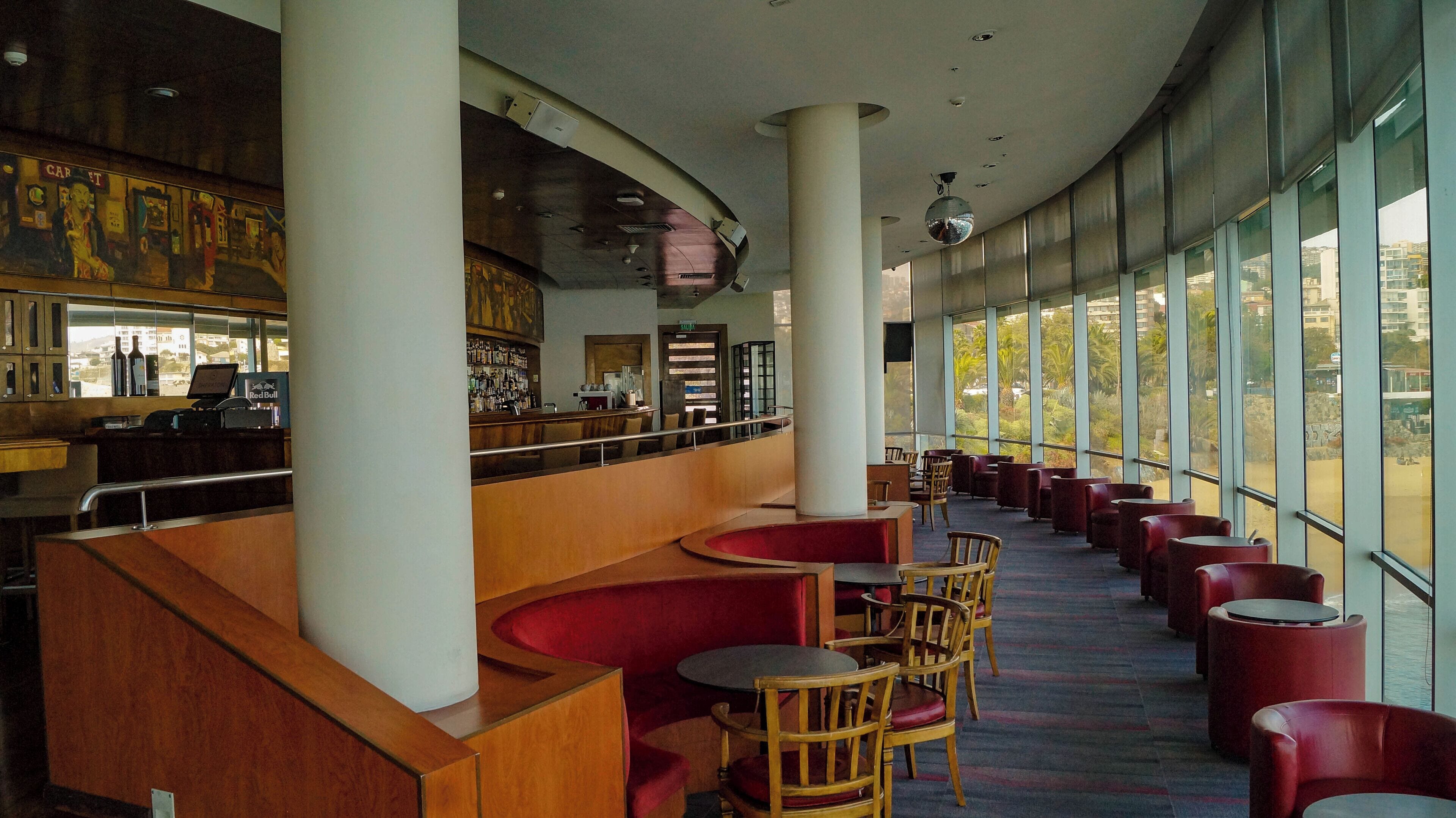 A spacious bar area with a curved counter, wooden chairs, and a view of the city through large windows.