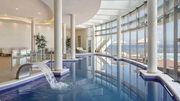 Un área de piscina interior moderna con vistas despejadas al océano, ventanas amplias y una cascada.