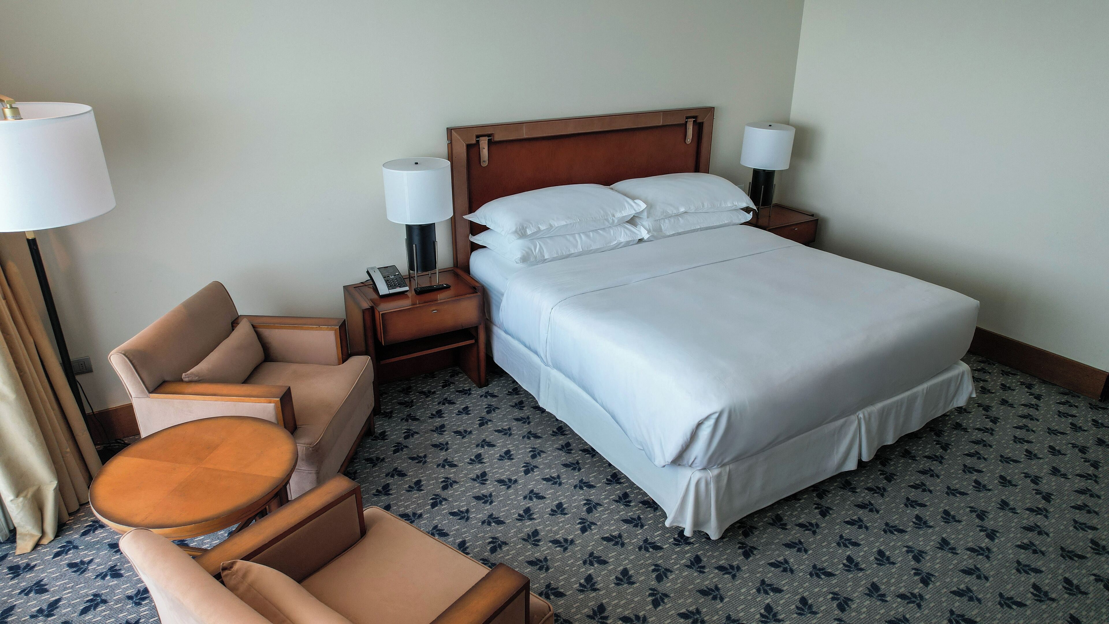 A hotel room with a bed, two bedside tables, a chair, a small table, and a lamp.