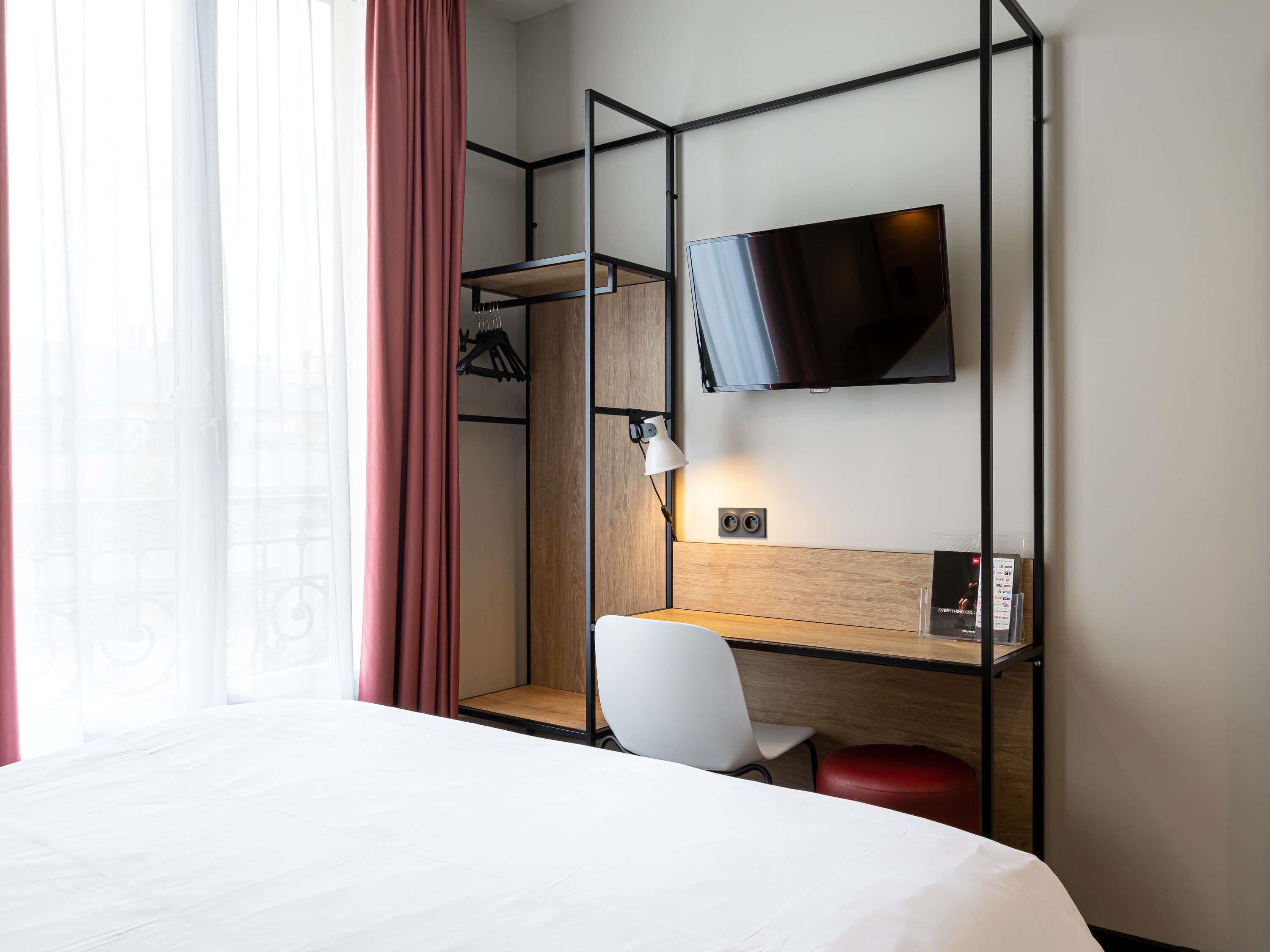 Een hotelkamer met een bed, een bureau met stoel, een aan de muur bevestigde televisie en een raam met gordijnen.