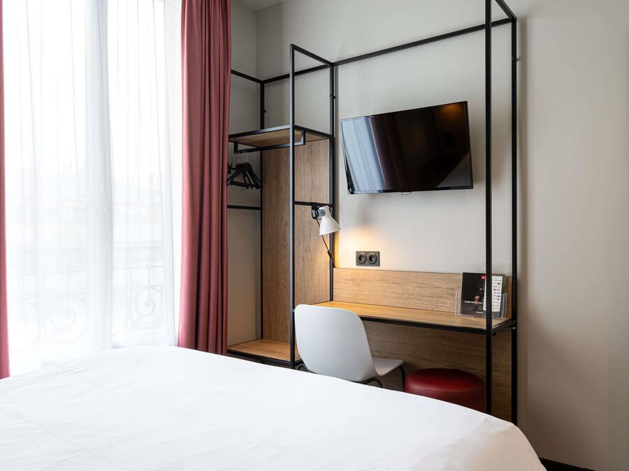 Een hotelkamer met een bed, een bureau met stoel, een aan de muur bevestigde televisie en een raam met gordijnen.