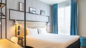 A modern hotel room with a large bed, a desk, and a chair. There are wall-mounted lamps and a telephone on the wall. - ibis Paris Boulogne Billancourt (Boulogne-Billancourt)