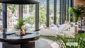 A person relaxing on a sofa with a view of a cityscape and mountains through large windows. - Hotel Therme Meran - Terme Merano (Merano)