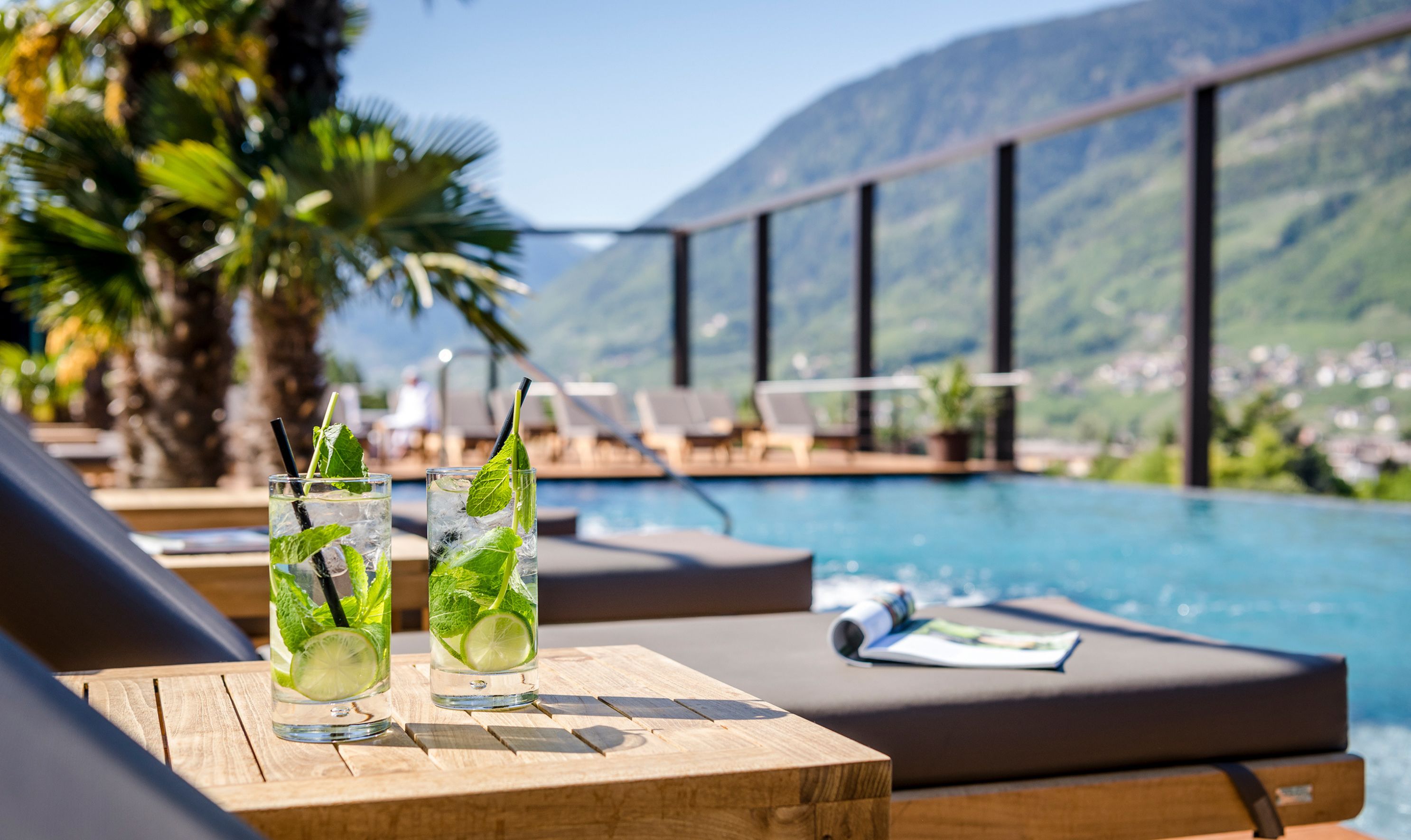 Two glasses of a refreshing beverage with mint leaves and lime slices on a wooden table by a poolside lounge area, overlooking a mountainous landscape.