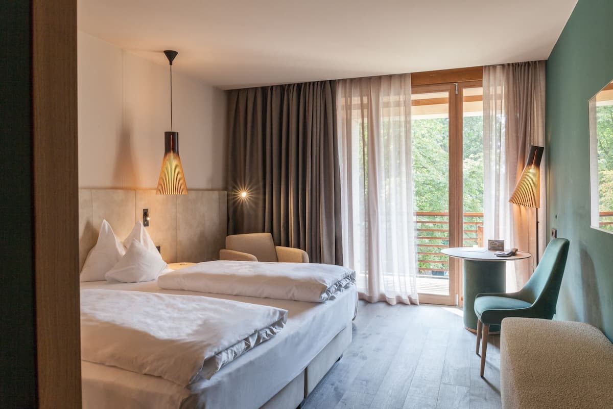 A hotel room with two beds, a desk, a chair, and a balcony with curtains.