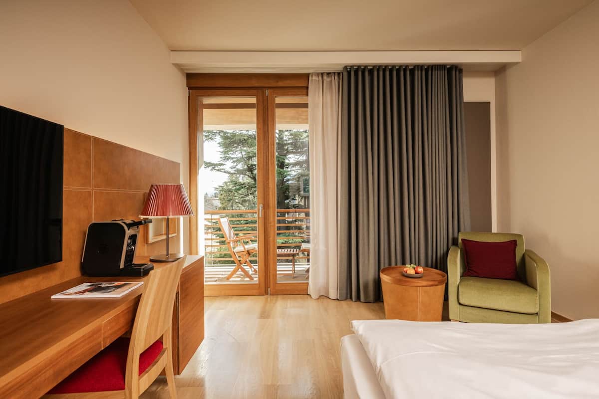 A hotel room with a bed, a desk with a chair, a green armchair, a small wooden side table, and a view of the outdoors through a sliding glass door.