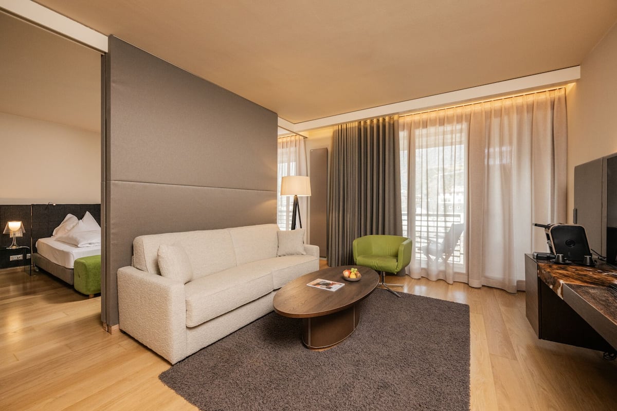 A modern hotel room with a sofa, a green armchair, a wooden coffee table, a desk with a computer, and a large window with curtains.