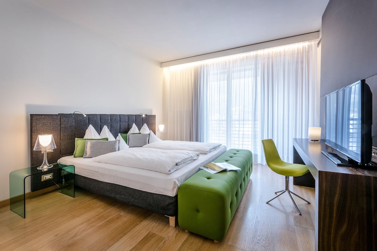 A modern hotel room with a bed, a green ottoman, a TV, and a nightstand with a lamp.
