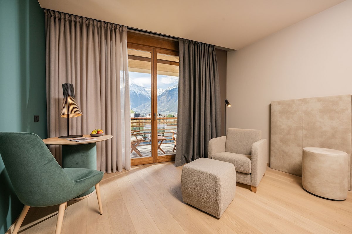 A modern hotel room with a wooden floor, a green armchair, a beige sofa, a small ottoman, and a table with a lamp and fruit bowl.
