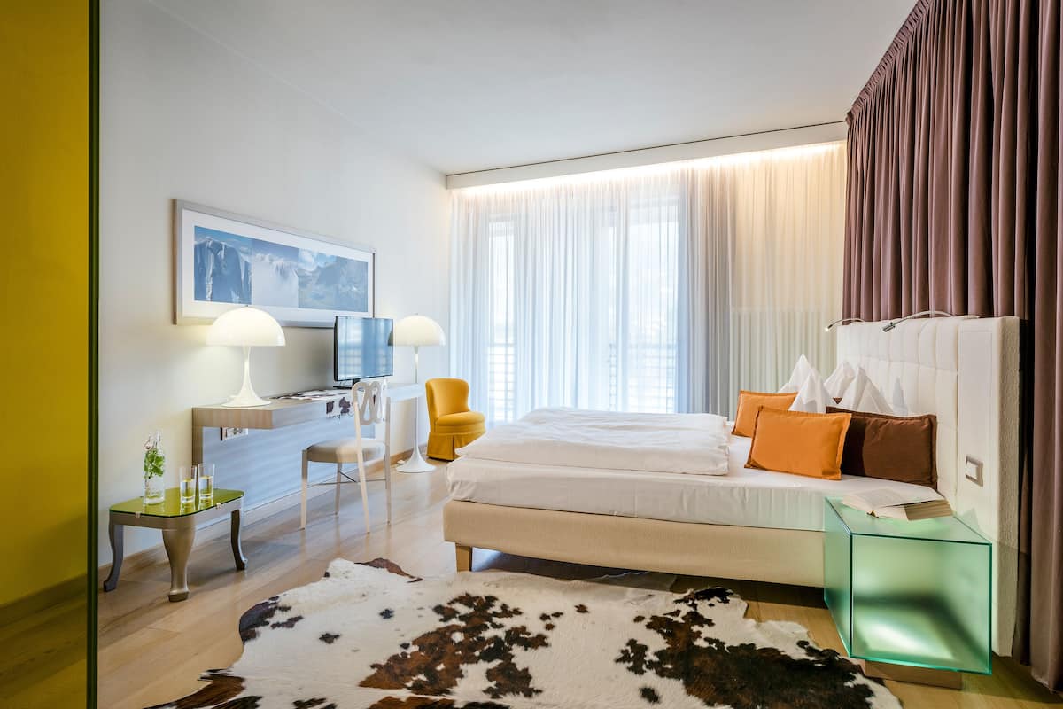 A modern hotel room with a bed, a desk, a chair, a television, and a large window with curtains.
