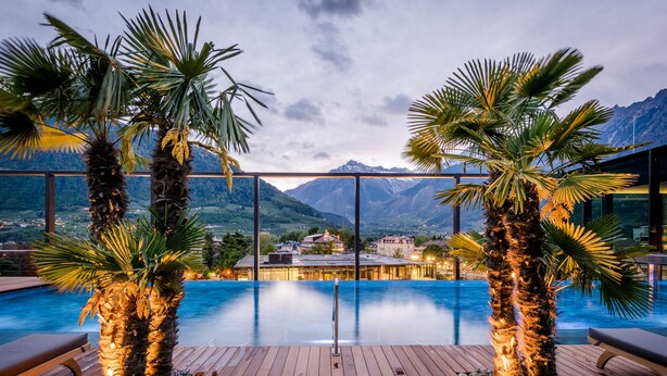 An infinity pool with wooden deck, palm trees, and a mountain view. - Hotel Therme Meran - Terme Merano (Merano)