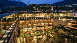 A modern hotel with a glass atrium, outdoor seating, and a view of the surrounding town and mountains. - Hotel Therme Meran - Terme Merano (Merano)