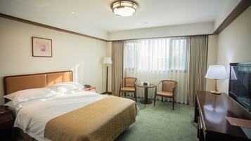 Habitación de hotel con una cama grande, dos sillas, un escritorio con televisor y una ventana con cortinas.