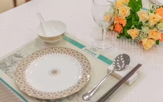 Ein gedeckter Tisch mit einem weißen Teller, einer Schüssel, einem Glas, einem Löffel und Essstäbchen, dazu ein Blumenstrauß.