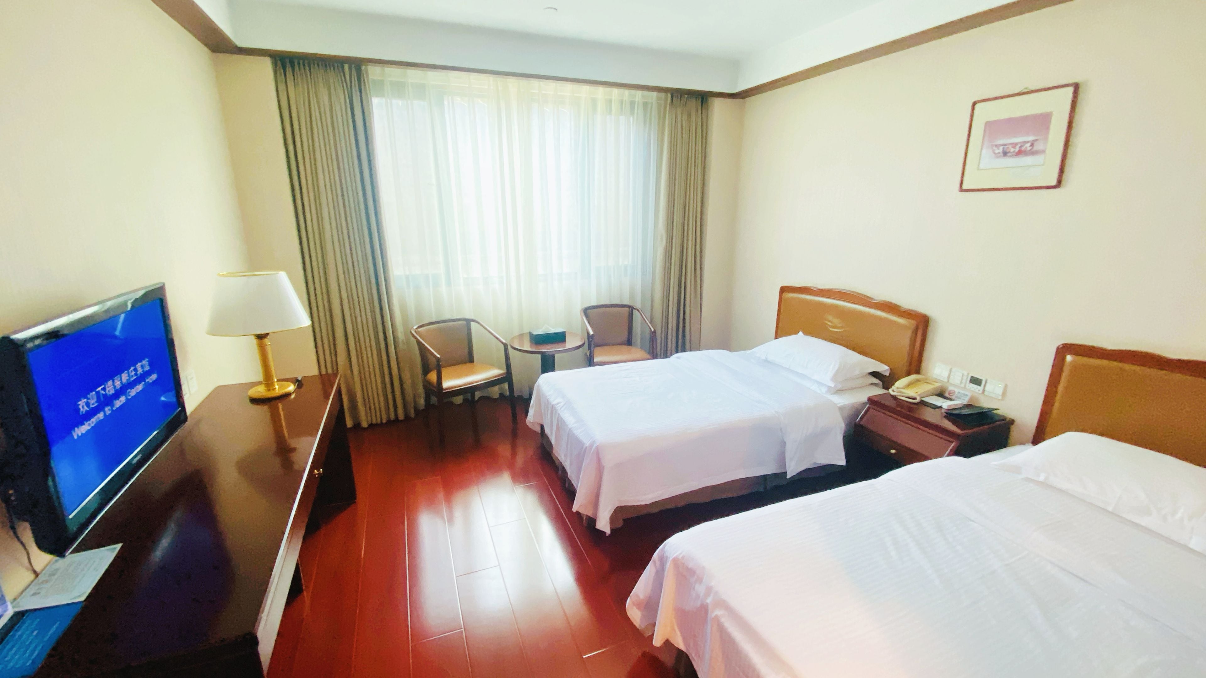 Habitación de hotel con dos camas, televisión, escritorio con lámpara, una silla y ventana con cortinas.