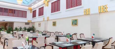 A spacious dining area with checkered chairs and tables, a high ceiling with a skylight, and traditional red lattice windows.