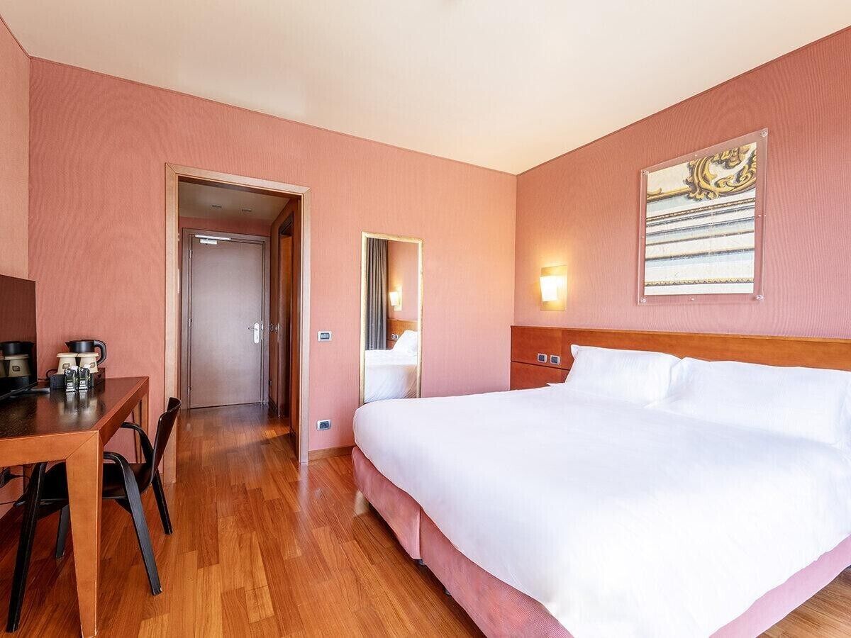 Une chambre d’hôtel avec un plancher en bois, un lit avec du linge de maison blanc, un bureau avec une chaise, un miroir et un luminaire fixé au mur.