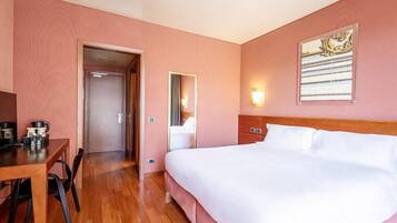 Une chambre d’hôtel avec un plancher en bois, un lit avec du linge de maison blanc, un bureau avec une chaise, un miroir et un luminaire fixé au mur.