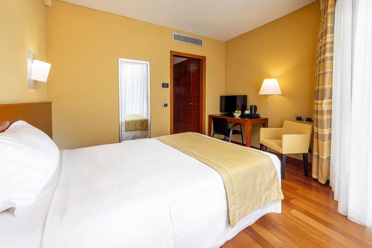 A hotel room with a large bed, a desk with a chair, a television, and a lamp.