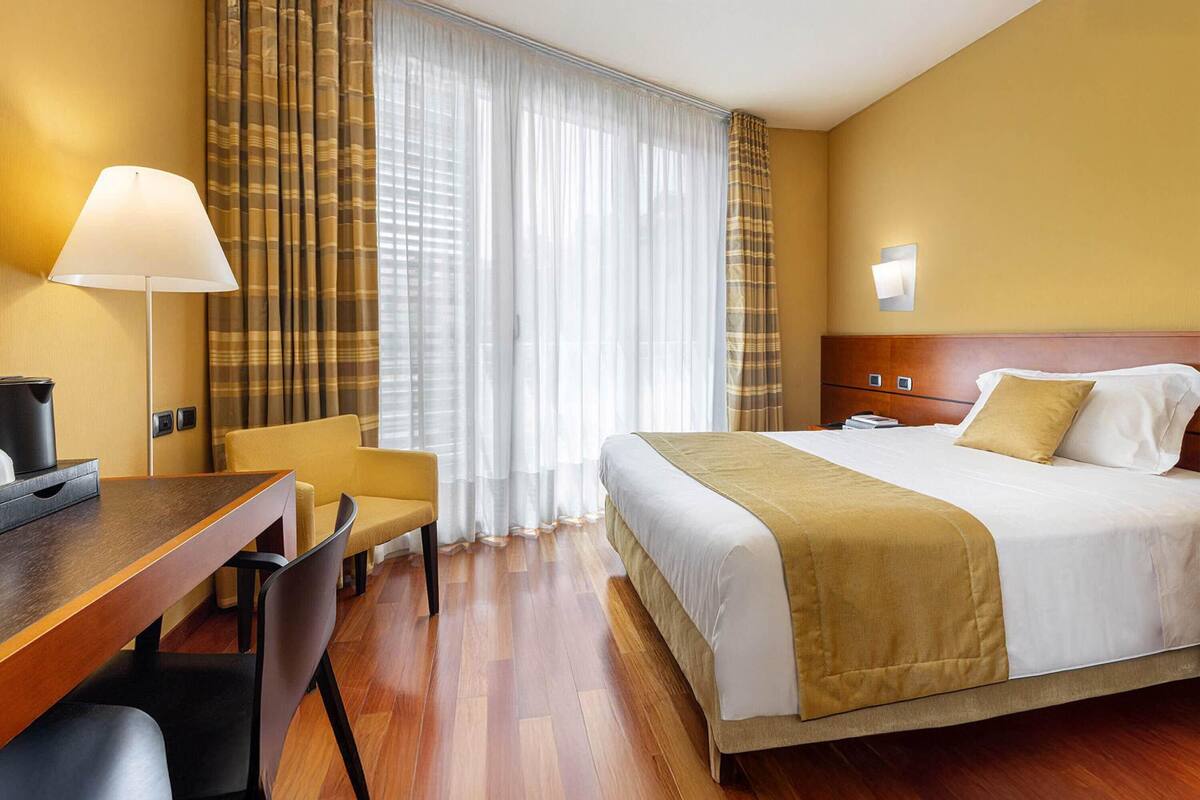 A hotel room with a bed, a desk, a chair, a lamp, and a window with curtains.