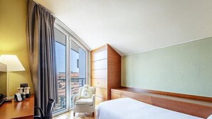 Habitación de hotel con cama, escritorio, silla, lámpara y vista de la ciudad a través de un ventanal grande.