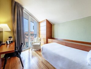 Habitación de hotel con cama, escritorio, silla, lámpara y vista de la ciudad a través de un ventanal grande.
