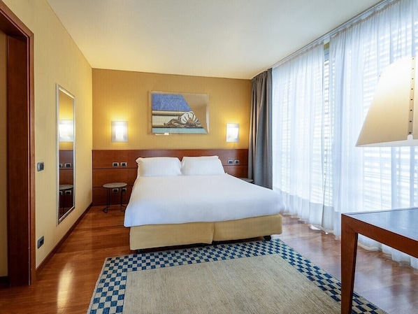 Habitación de hotel con una cama grande, una mesita pequeña, una silla, una lámpara de pared y un cuadro.