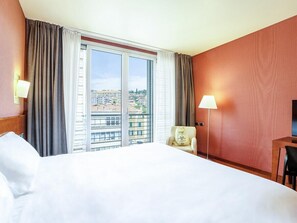 A hotel room with a large bed, a chair, a desk, and a view of the city through the window. - B&B HOTEL Imperia Rossini al Teatro (Imperia)