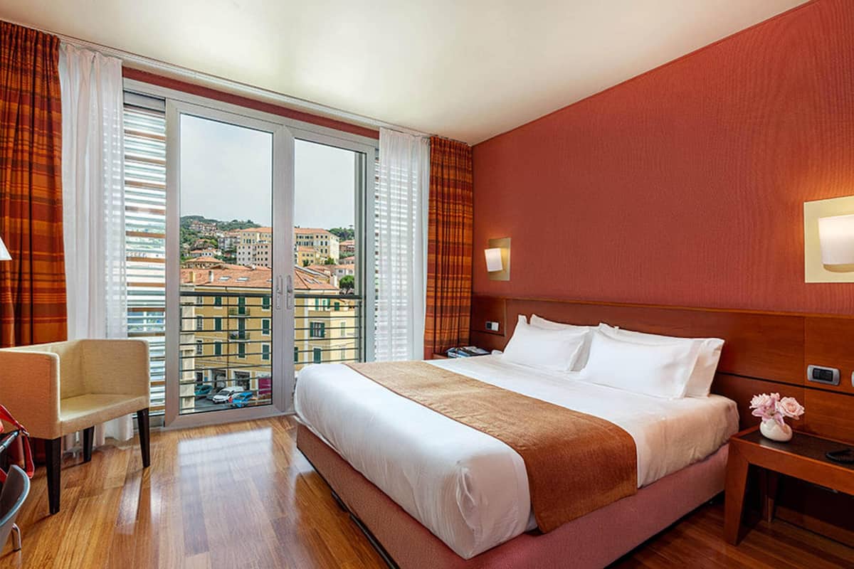 A hotel room with a large bed, a desk, a chair, a nightstand, and a view of buildings through a sliding glass door.