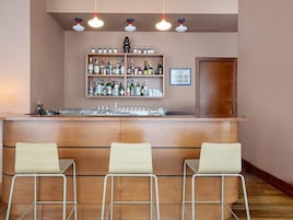 Zona de bar con mostrador de madera, tres taburetes y estante con diferentes botellas y vasos.