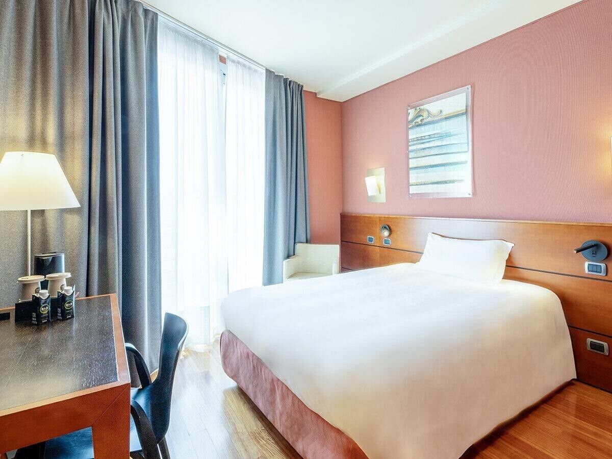 A hotel room with a bed, a desk with a lamp, a chair, and a window with curtains.