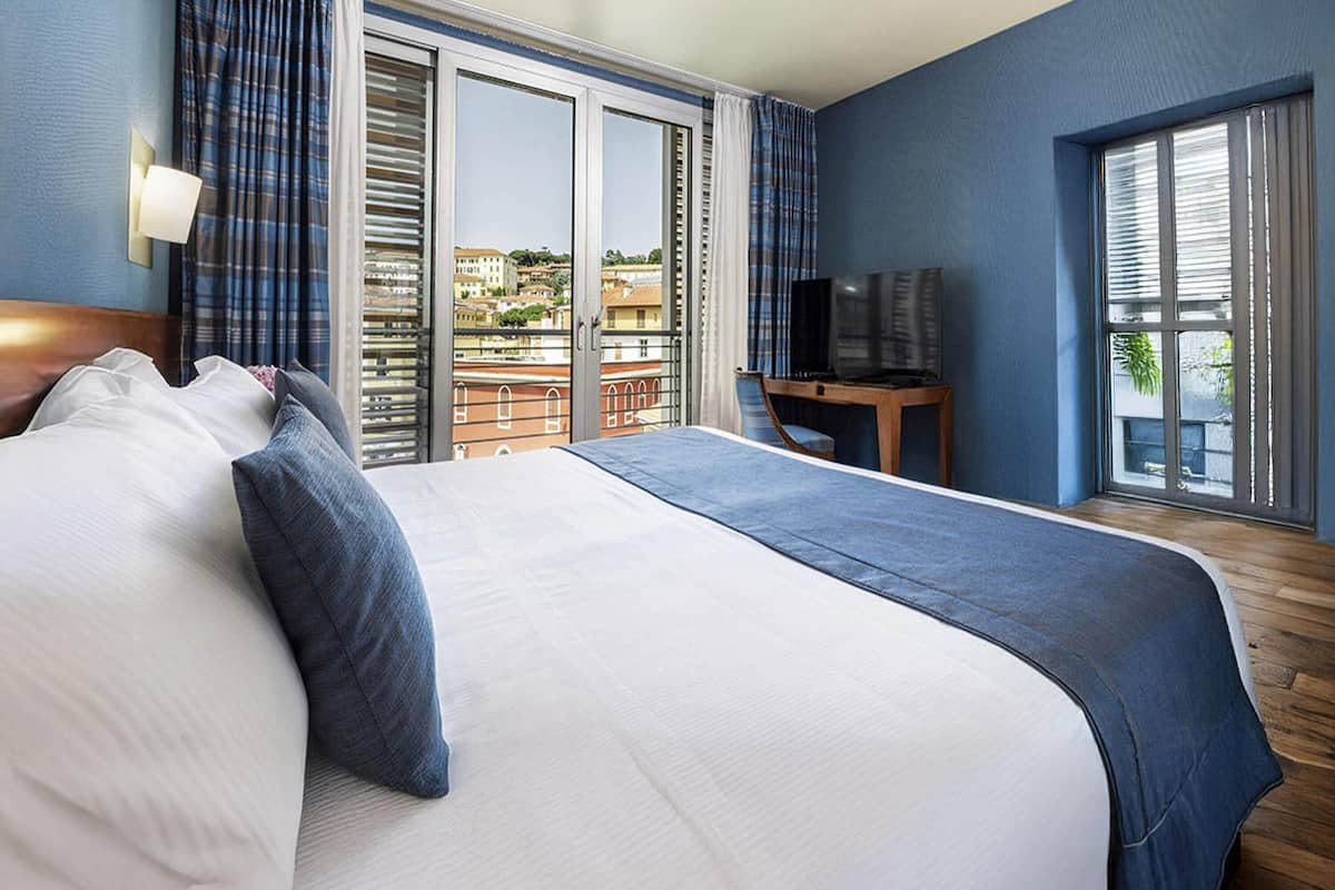 A hotel room with a large bed, a desk with a TV, and a balcony with a city view.