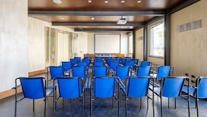 A conference room with rows of blue chairs, a projector screen, and large windows. - B&B HOTEL Imperia Rossini al Teatro (Imperia)