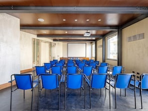 A conference room with rows of blue chairs, a projector screen, and large windows. - B&B HOTEL Imperia Rossini al Teatro (Imperia)