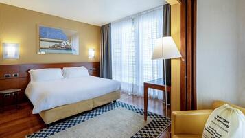 Quarto de hotel com cama, mesa de cabeceira, luminária, cadeira e uma bolsa decorativa.