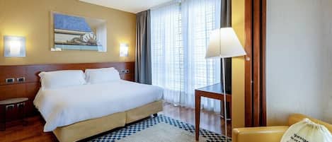 Una habitación de hotel con cama, mesita de noche, lámpara, silla y un bolso decorativo.