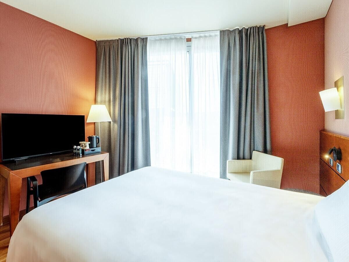 A hotel room with a bed, a desk with a television, a chair, and a lamp.
